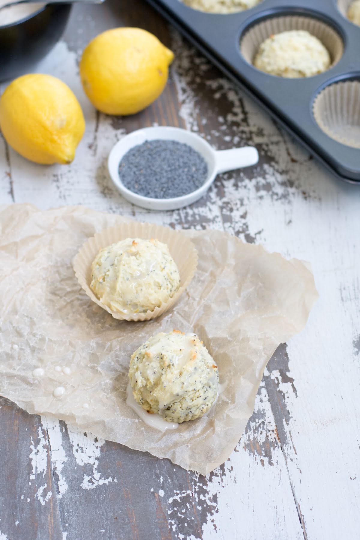 These are the perfect Gluten Free Lemon Poppyseed Muffins. Real lemon and crunchy poppy seeds in a tender, sweet muffin. Easy to make! Two small muffins on a piece of parchment, next to a dish of poppy and two lemons.