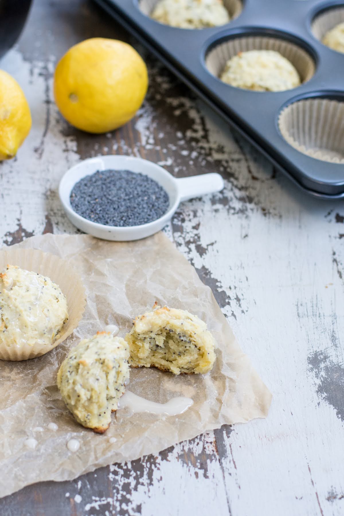 These are the perfect Gluten Free Lemon Poppyseed Muffins. Real lemon and crunchy poppy seeds in a tender, sweet muffin. Easy to make! Muffins in a tin, with two removed from the muffin tin and placed on the counter. A dish of poppy seeds and two lemons are also on the counter.