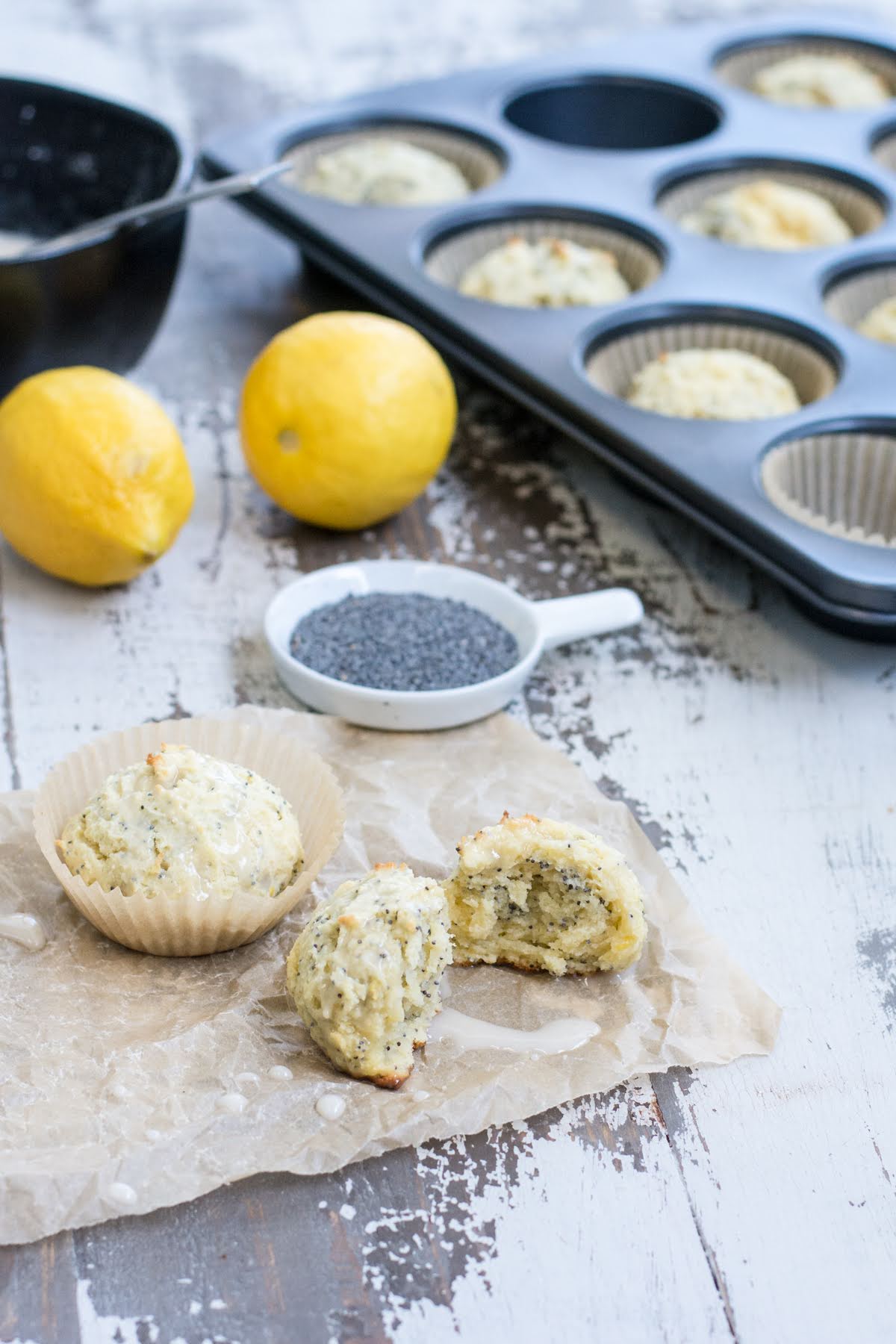 These are the perfect Gluten Free Lemon Poppyseed Muffins. Real lemon and crunchy poppy seeds in a tender, sweet muffin. Easy to make! A muffin tin with muffins resting inside, next to a bowl of glaze, some lemons, poppy seeds, and two additional muffins. One of the loose muffins has been broken in half.