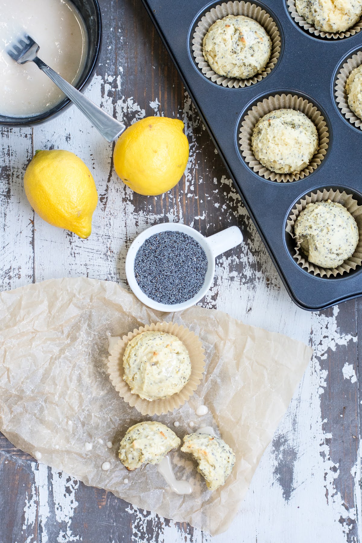 These are the perfect Gluten Free Lemon Poppyseed Muffins. Real lemon and crunchy poppy seeds in a tender, sweet muffin. Easy to make! Overhead shot of a table with a muffin tin, lemons, glaze, and other items. Two muffins are nearby on the counter.
